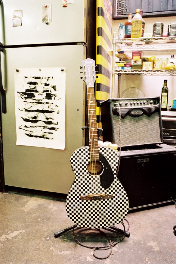 Eine schwarz-weiß-karierte Gitarre auf einem Gitarrenständer steht vor einem Kühlschrank und einem Verstärker