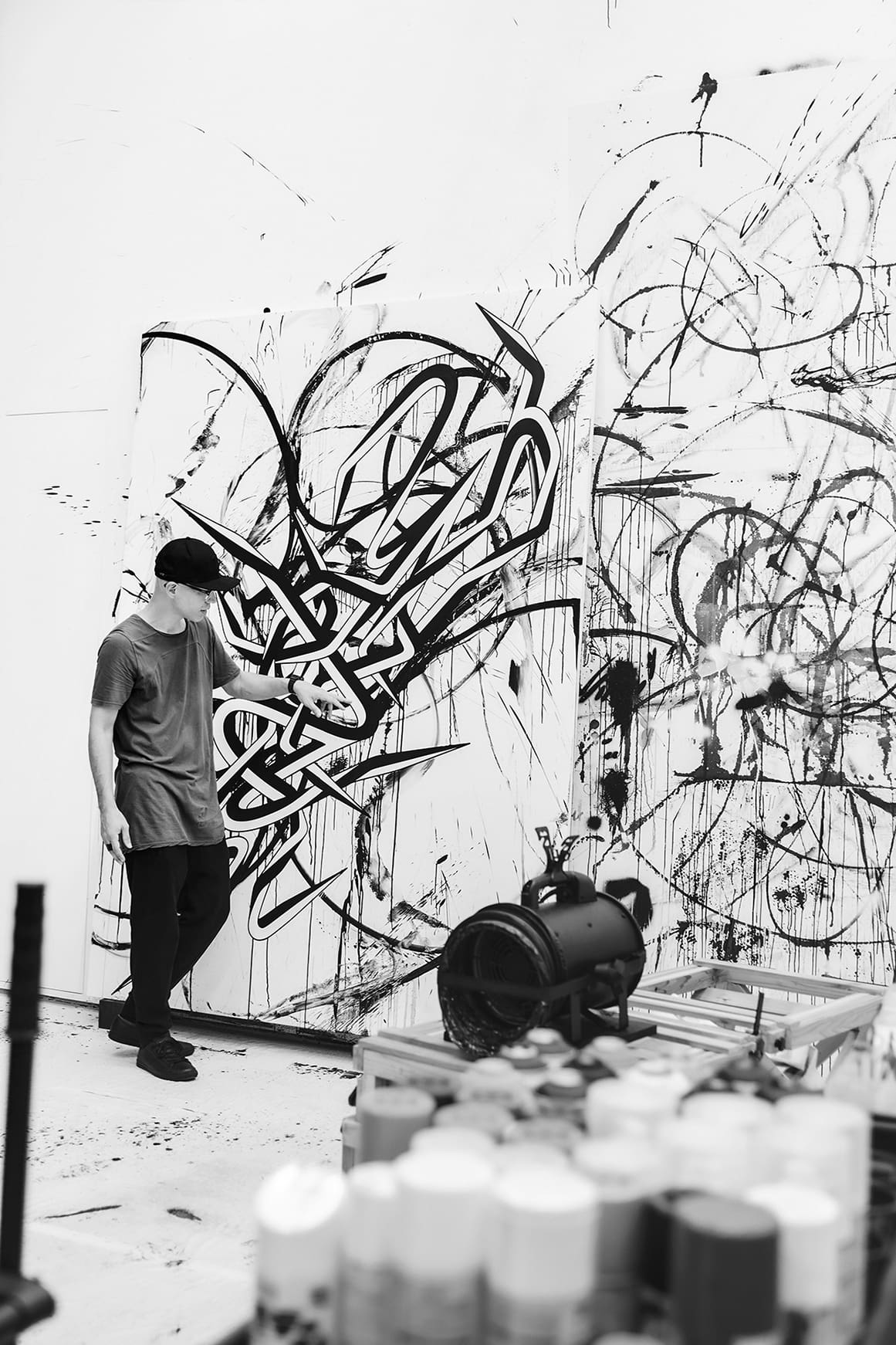 Der Künstler Enrico Isamu Oyama steht vor einer weißen Wand an der schwarze Graffitizeichnungen zu sehen sind