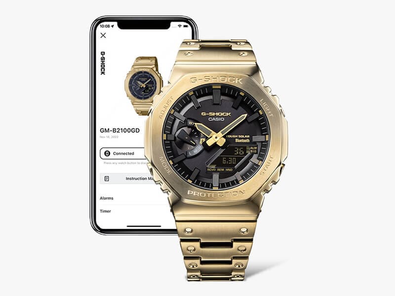 Eine Casio G-Shock Vollmetalluhr mit schwarzem Zifferblatt sowie goldfarbenem Armband und Gehäuse vor einem Smartphone, auf dessen Display Informationen zur Uhr zu sehen sind
