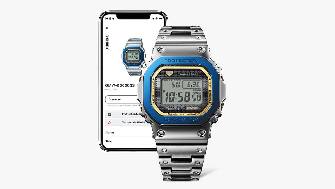 Die Casio G-Shock GMW-B5000SS-2 Jubiläumsarmbanduhr mit silbernem Edelstahlarmband, blauer Lünette und goldenem Zifferblattrand vor einem Smartphone, auf dessen Display Informationen zur Uhr zu sehen sind