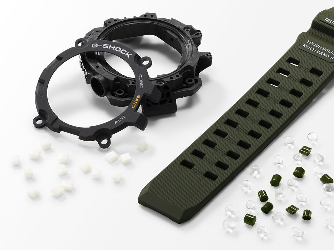 Ein Gehäuseteil, eine Lünette und ein Armband umgeben von kleinen durchsichtigen und schwarzen Resingranulat-Teilchen