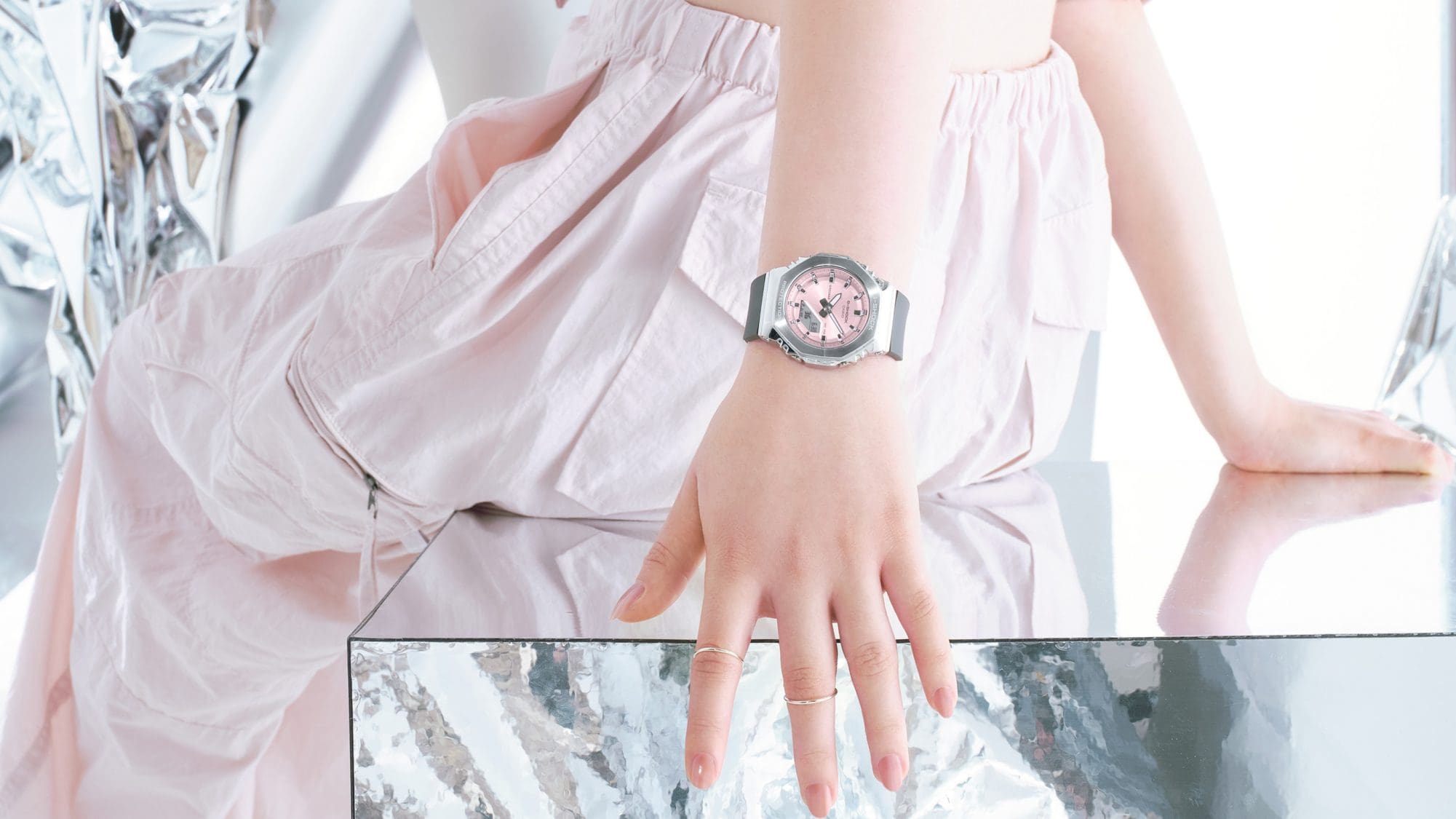 Die Casio G-Shock GM-S2110-4A Armbanduhr mit rosanem Zifferblatt, silberfarbenem Gehäuse und schwarzem Armband am Handgelenk einer Frau, die auf einem verspiegeltem Hocker sitzt