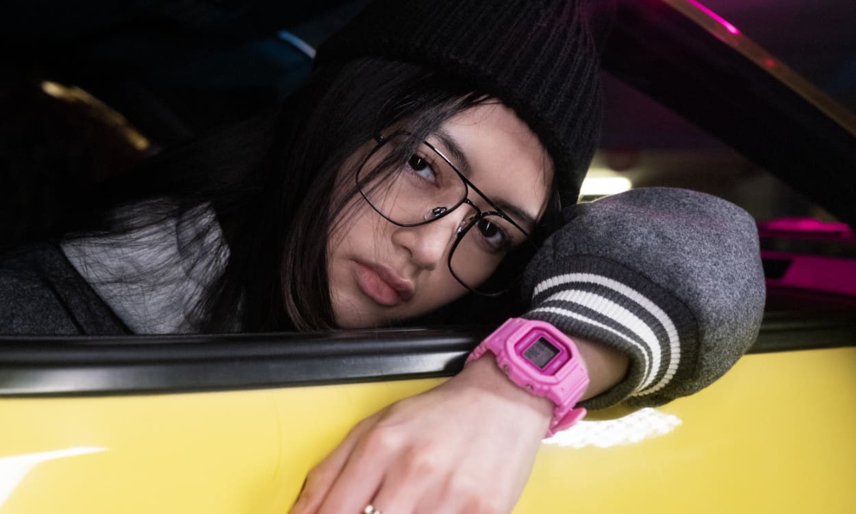 Eine Frau lehnt sich vom Beifahrersitz aus einem gelben Auto, trägt einen schwarzen Hut, eine Brille und die pinkfarbene Digitaluhr GMD-S5600PP von G-SHOCK.