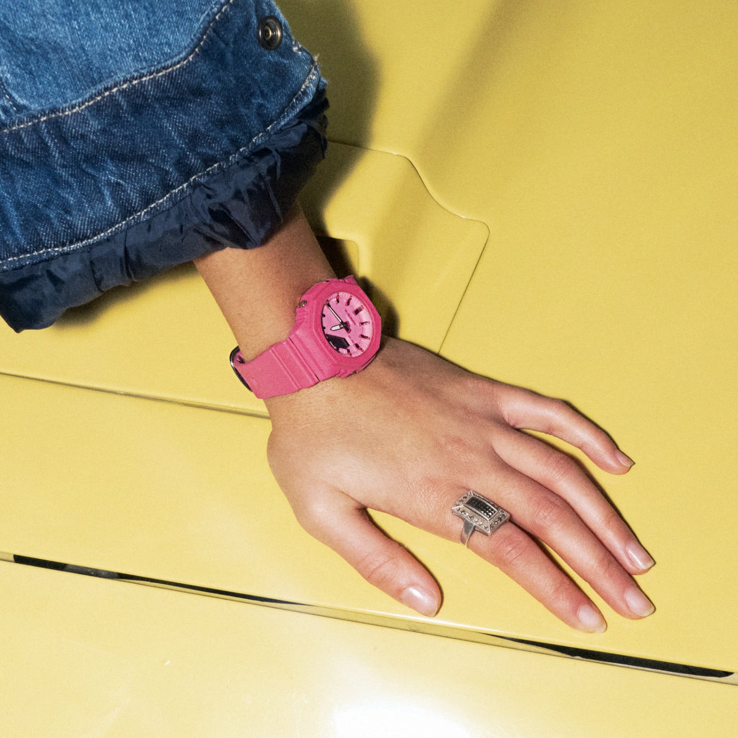 Eine Frauenhand, die sich auf eine gelbe Motorhaube stützt und die rosa Analoguhr GMA-2100PP-4 trägt.