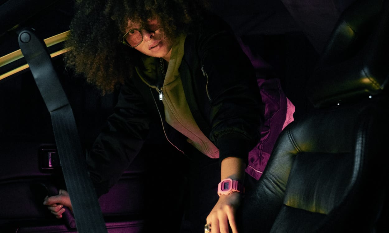 Eine Frau mit Afro, schwarzer Jacke und der rosa G-SHOCK GMD-S5610 lehnt sich auf den Beifahrersitz eines Autos.