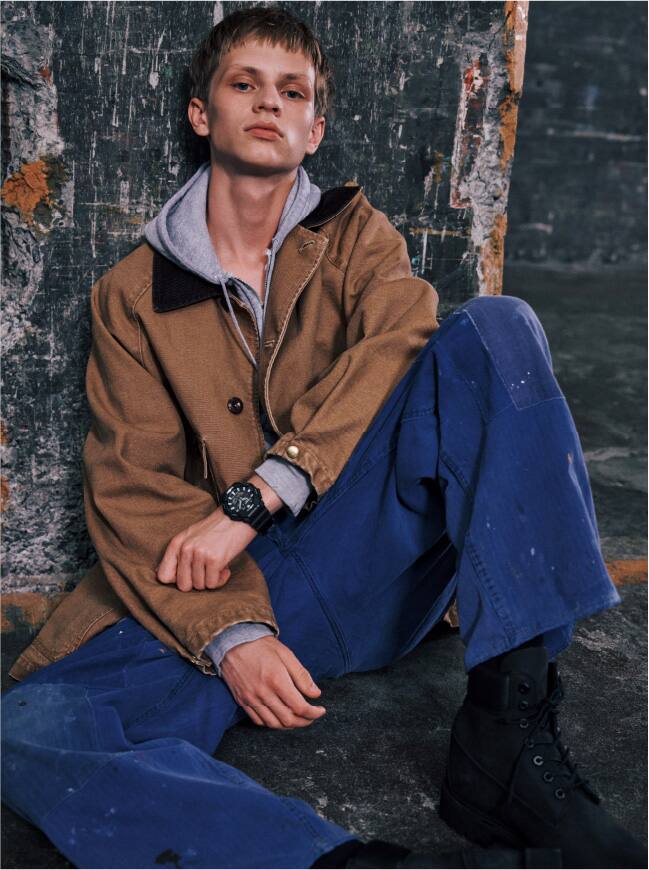 A person in beige/brown jacket an a blue jeans sitting in front of a wall wearing the black G-Shock GA-B010-1A in black with an analogue-digital display on his wrist.