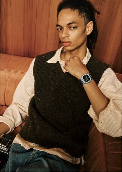 A man with dreadlocks, a beige shirt and a brown slipover wearing the G-Shock GM-5600M-1 on his wrist.