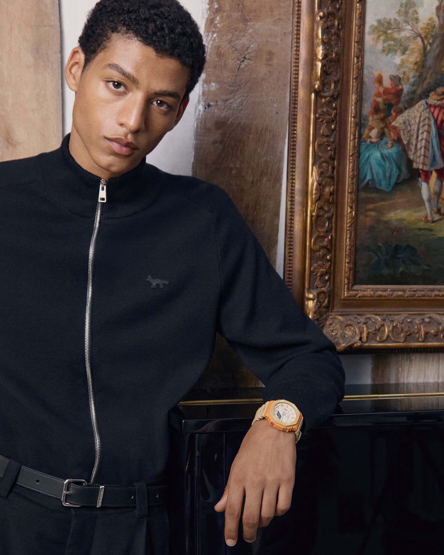 A man wearing a blue cardigan from Maison Kitsune and the beige/orange G-SHOCK GA-2100MK-5A on his wrist leans on a piano