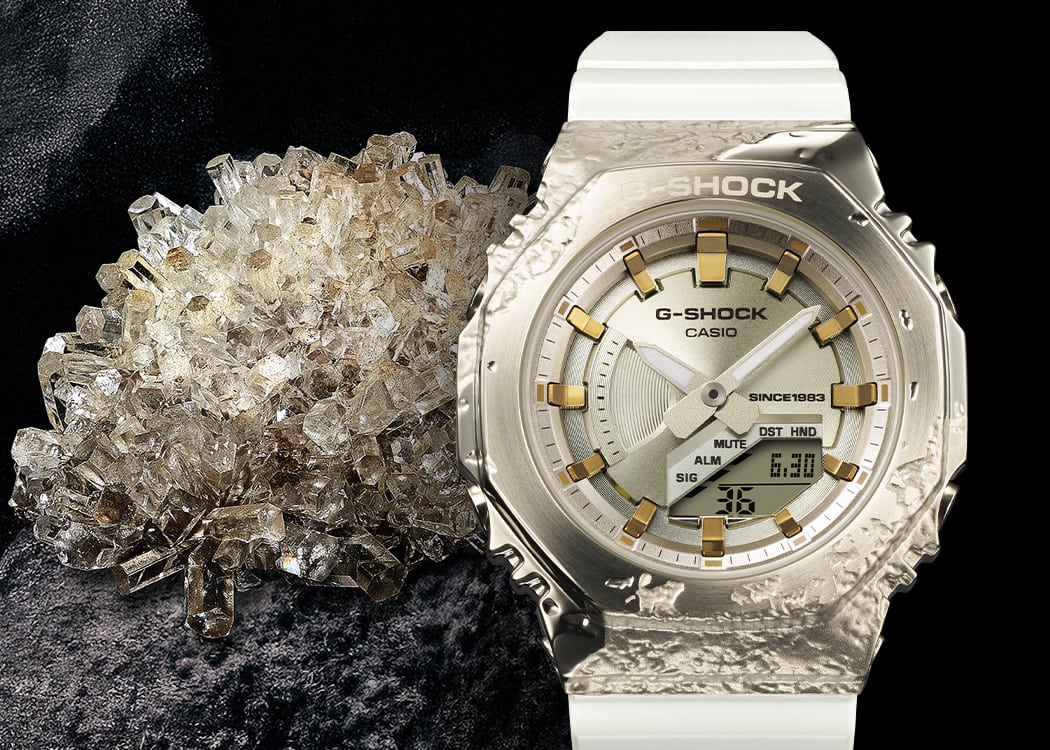GM-S2140GEM-9A El reloj aniversario GM-S2140GEM-9A, con un brillante acabado dorado y correa blanca, junto a cristales de calcita transparentes con reflejos dorados.