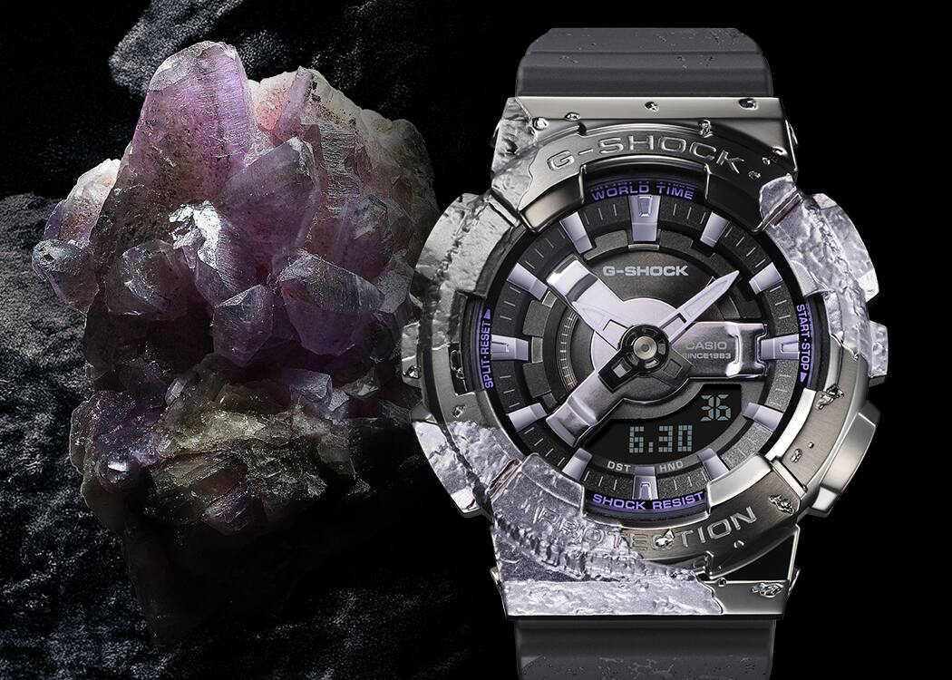GM-S114GEM-1A2 El reloj aniversario GM-S114GEM-1A2, de color gris oscuro brillante, junto a un cristal de calcita de color púrpura brillante.