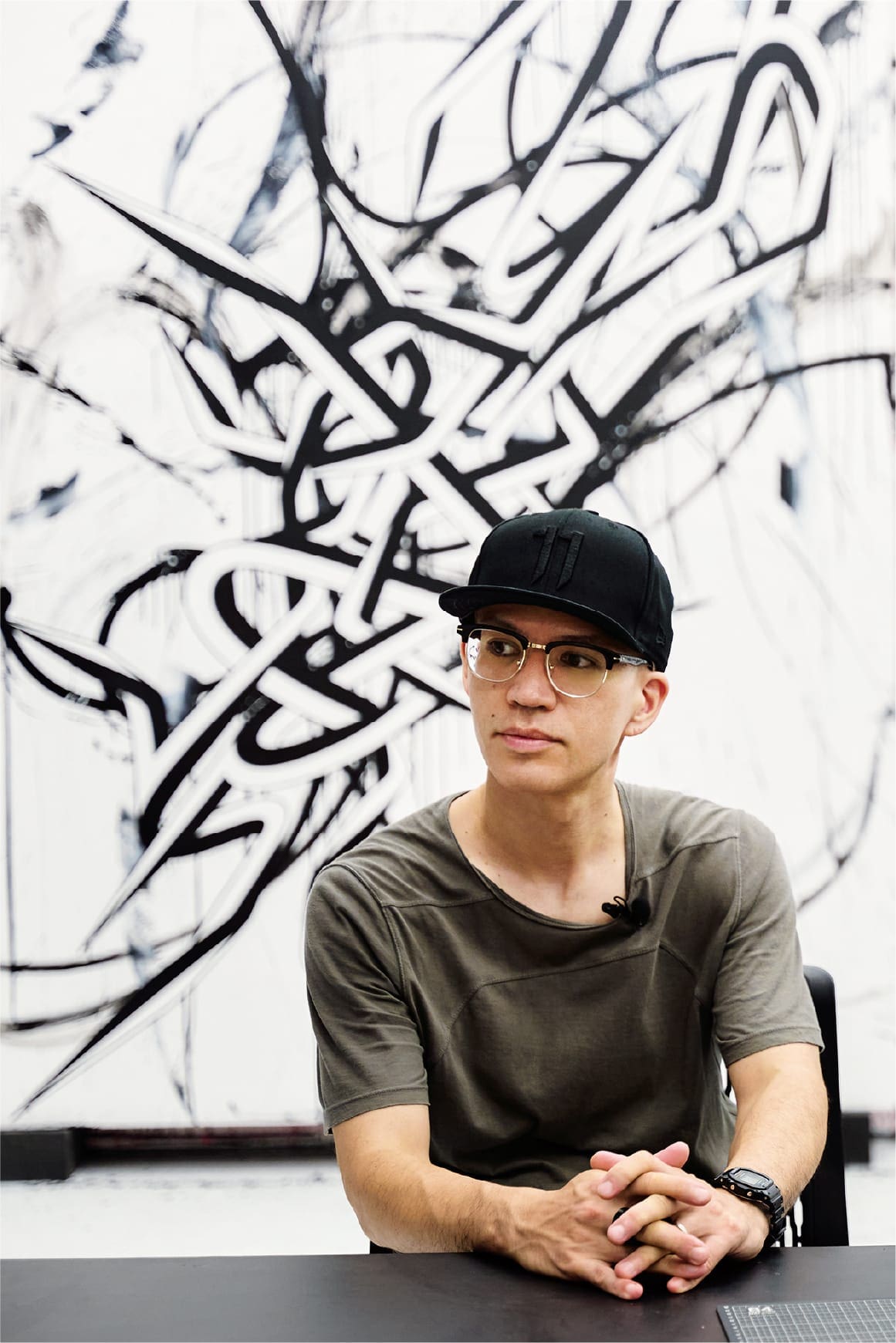 Enrico Isamu Oyama con gorra negra, camisa marrón y reloj digital G-SHOCK negro sentado frente a una de sus obras de arte.