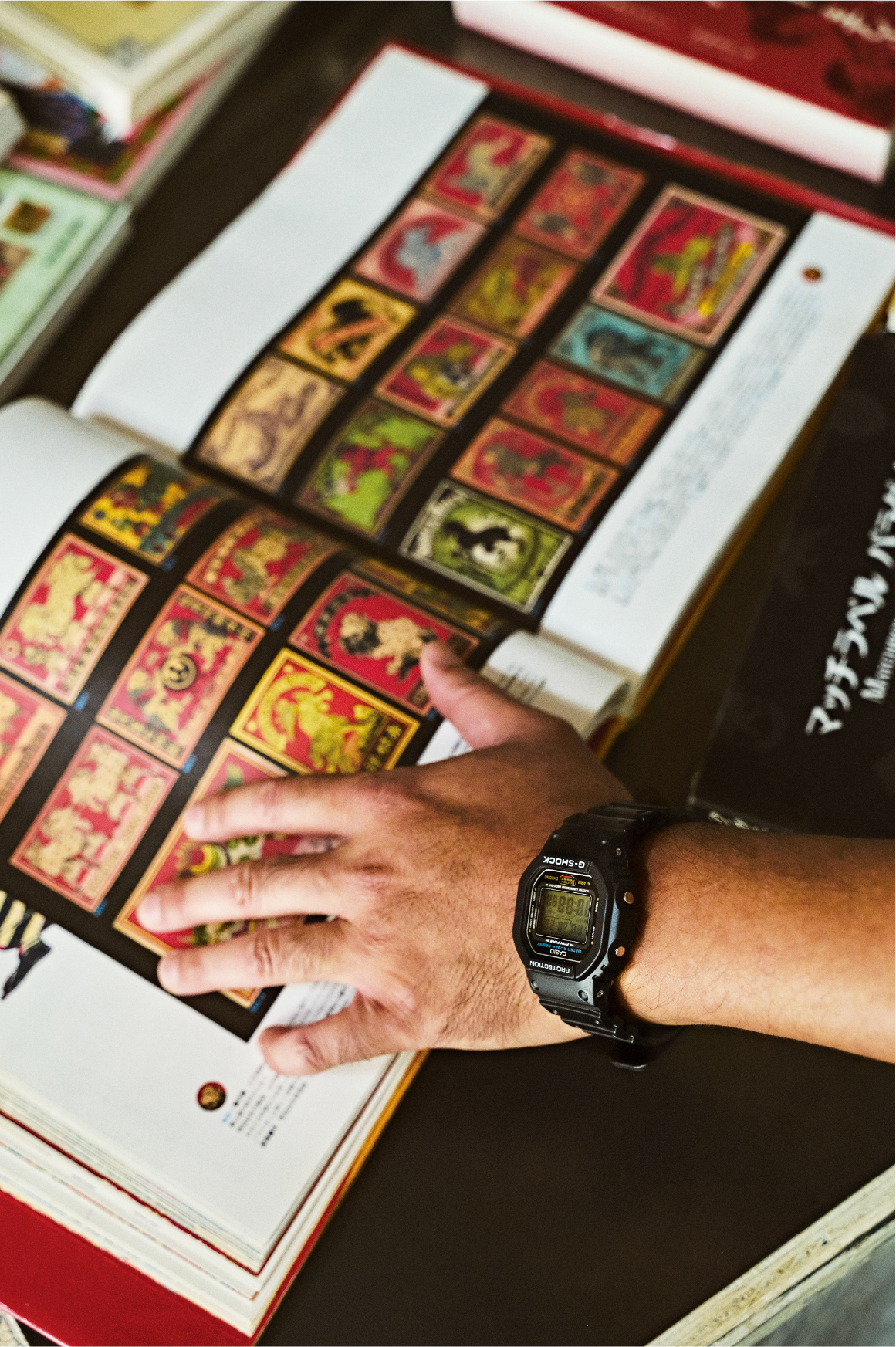Una mano acaricia una página de libro con diseños de cerillas retro, en la muñeca un reloj G-SHOCK.