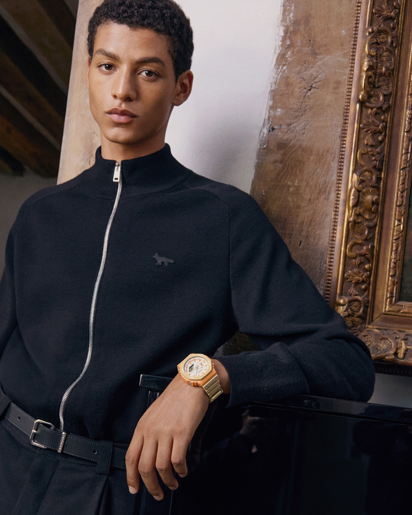 Un hombre con un cárdigan azul y el reloj de colaboración G-SHOCK & Maison Kitsune en la muñeca se apoya en un piano y mira directamente a la cámara