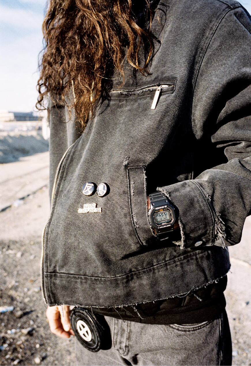 Une main dans la poche d'une veste en jean noir délavé. Une G-Shock noire apparaît entre la poche et la manche.