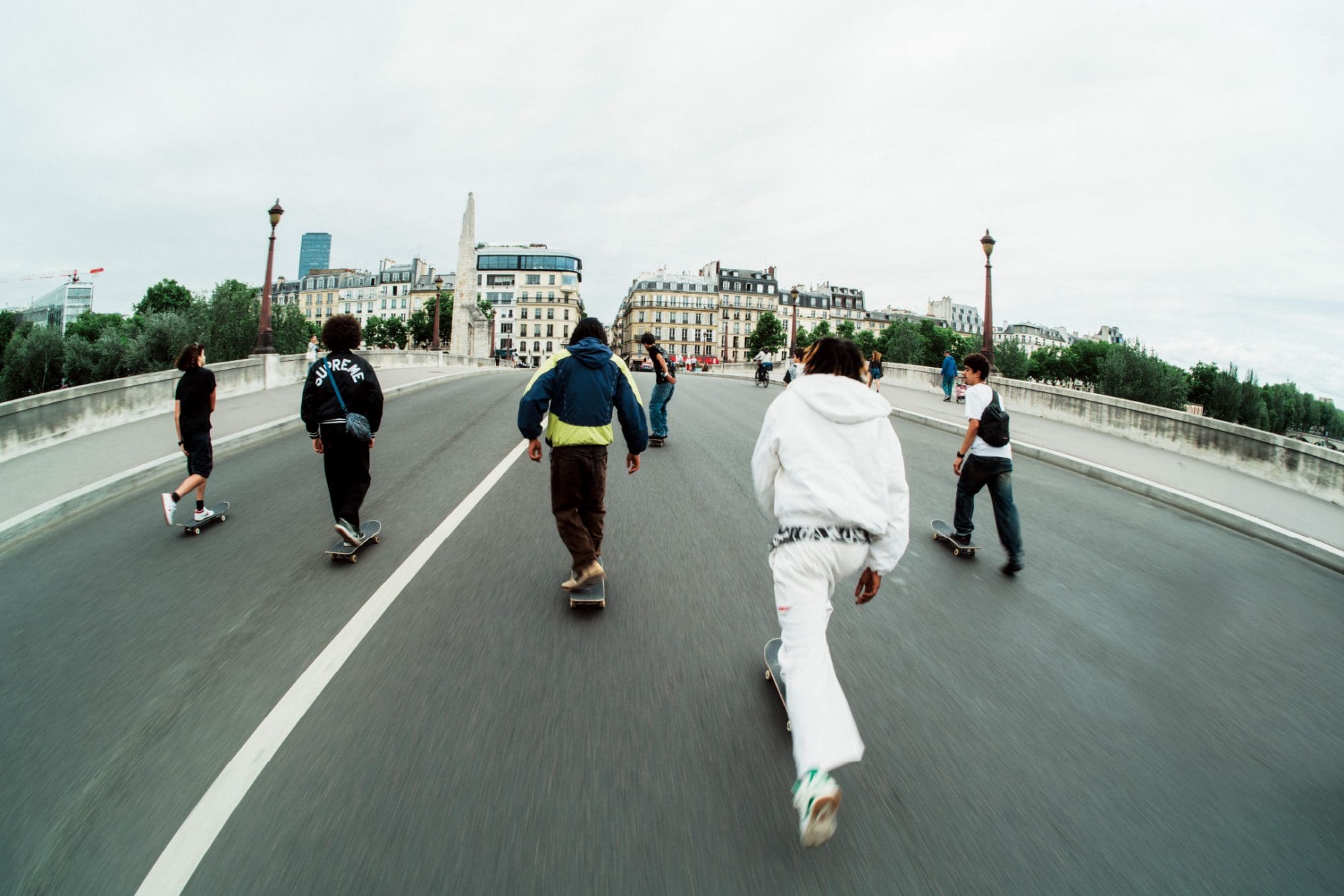 Groupe de skateurs traversant un pont à Paris avec la ligne d'horizon de la ville en arrière-plan.