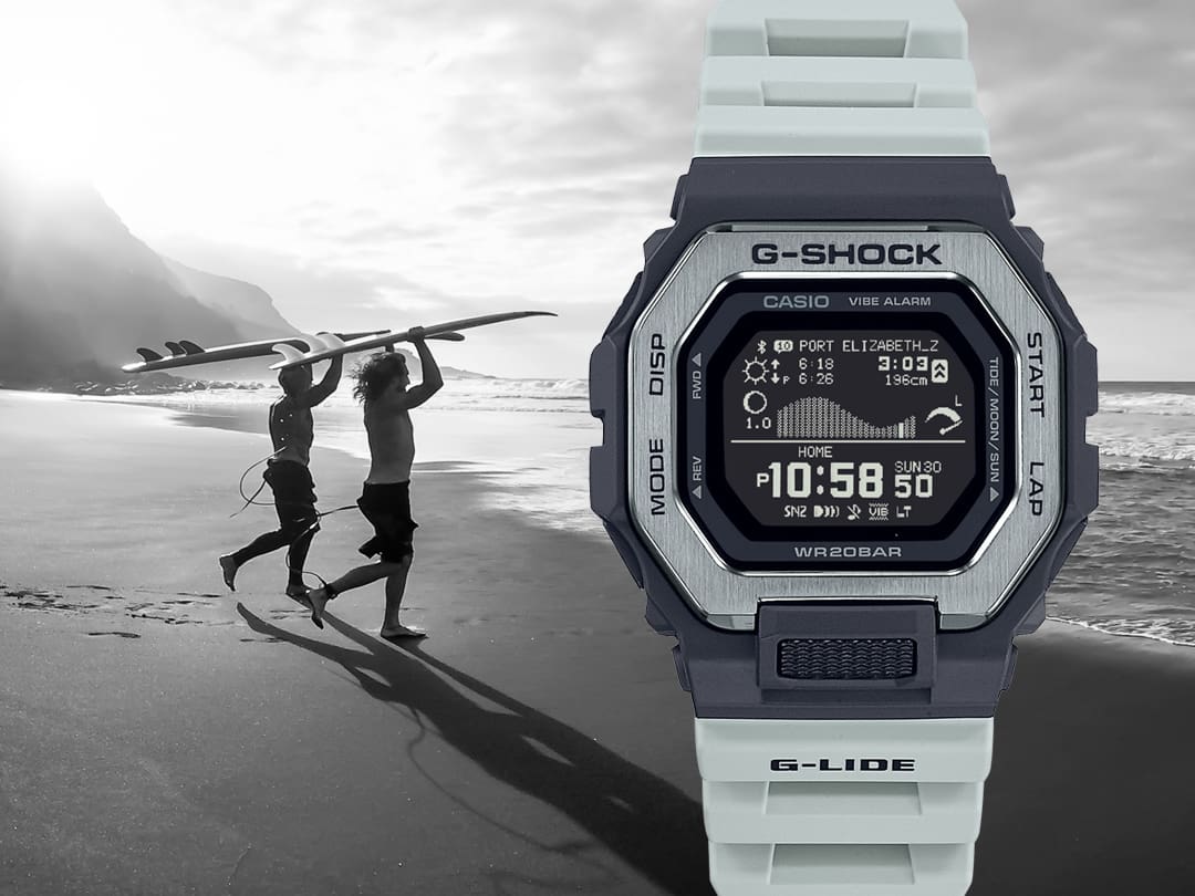 Devant : la montre Casio G-Shock GBX-100TT-8 avec un bracelet en plastique gris clair, un boîtier noir et une lunette argentée, derrière : une photo en noir et blanc de deux surfeurs courant sur la plage vers la mer.