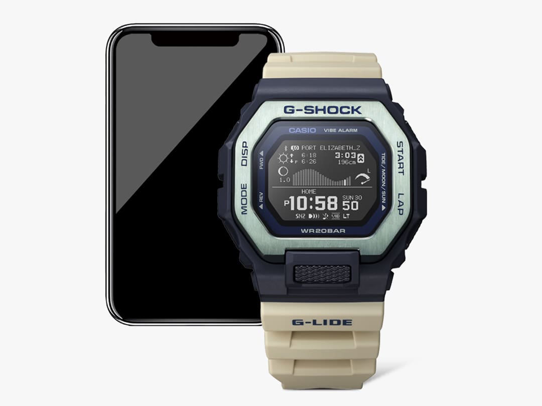 Devant : une montre surf Casio GBX-100-2 avec un bracelet beige, un boîtier noir et une lunette en acier inoxydable argenté, derrière : Un smartphone affiche des informations sur le spot de surf de Port Elizabeth en Afrique du Sud dans l'application G-Shock Move.