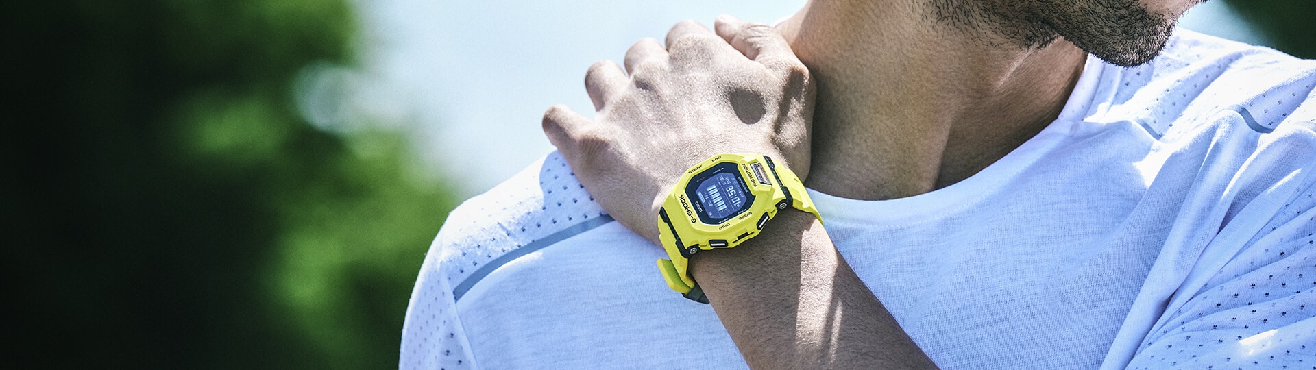 Un homme en t-shirt de sport blanc se saisit de l'épaule droite avec la main gauche, il porte au poignet une montre-bracelet Casio Shock GDB-200 jaune.