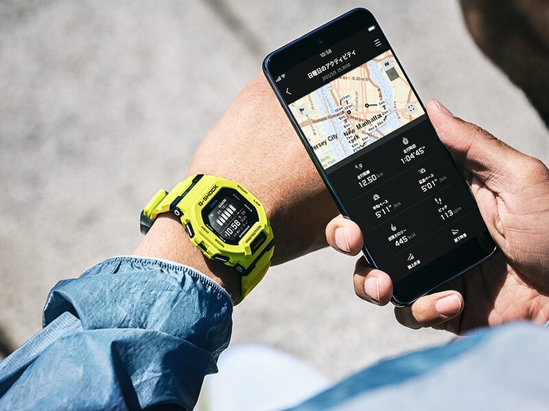 Quelqu'un regarde sa montre G-SQUAD GBD-200-9 en tenant son smartphone à côté de la montre, sur laquelle l'application G-Shock Move affiche, entre autres, les pas effectués, les kilomètres parcourus et les calories dépensées.