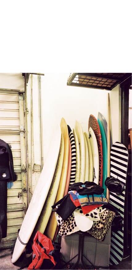 Più di una dozzina di tavole da surf sono appoggiate a una parete. Vari tessuti sono accatastati su uno sgabello davanti a loro, e una tavola da snowboard poggia accanto a esse