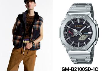 O relógio de metal GM-B2100 com um mostrador analógico preto, ao lado de um homem que usa o G-SHOCK e um colete axadrezado.