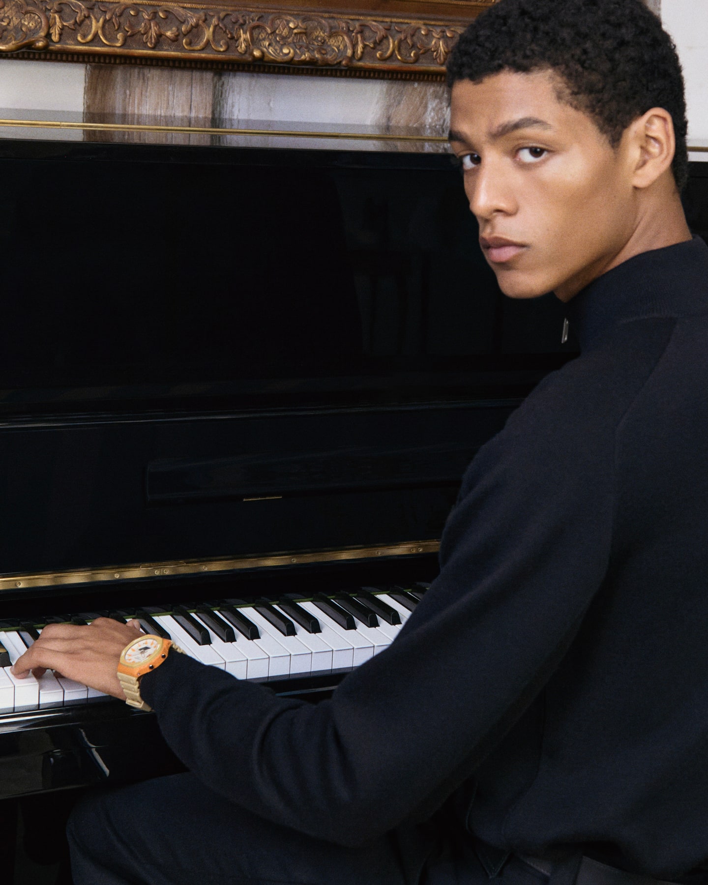Um homem com um casaco de malha azul e o relógio da colaboração G-SHOCK & Maison Kitsune no pulso senta-se num piano e olha diretamente para a câmara
