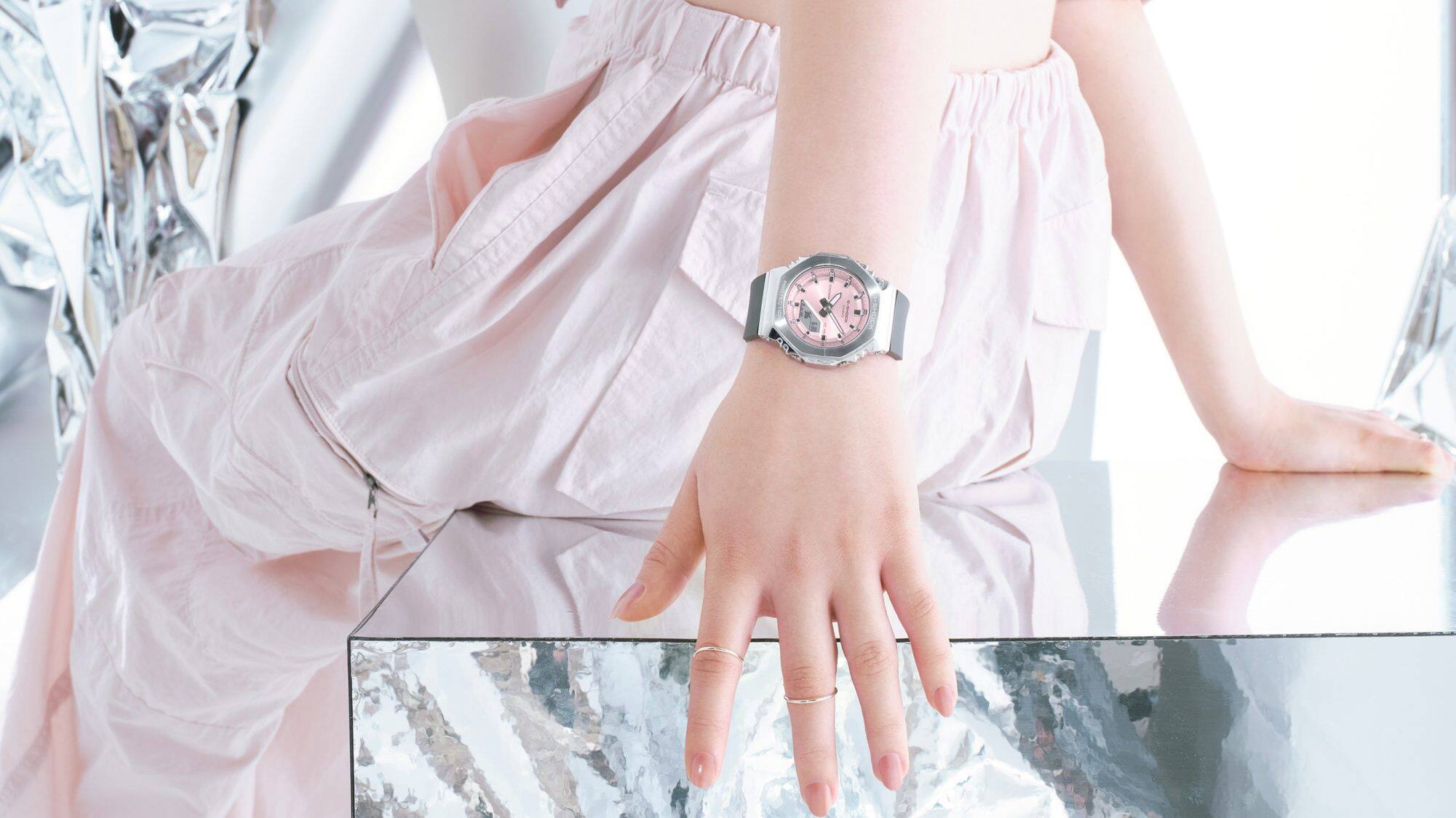 O relógio de pulso Casio G-Shock GM-S2110-4A com mostrador cor-de-rosa, caixa prateada e bracelete preta no pulso de uma mulher sentada num banco espelhado